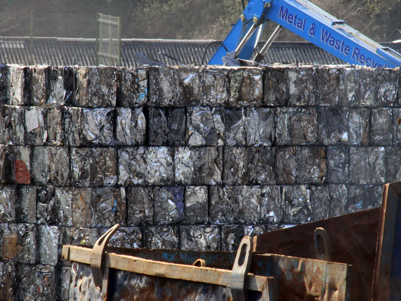 Mur de balles de ferraille compactées empilées sur un parc à ferrailles, avec bennes et grue de manutention